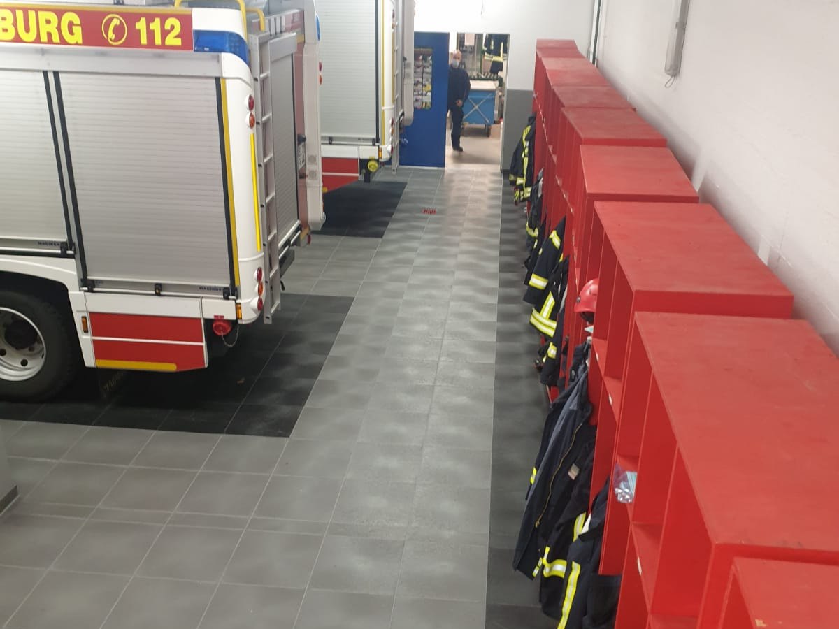 Feuerwache mit Fahrzeugen und roten Spinden, der Boden trennt Stellflächen und Gang in Schwarz und Grau